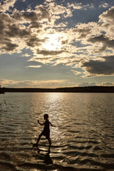 Naklejka na ściany i meble Silhouette of a boy walking on the water in the rays of the sunset. Beach by the sea, lake or river