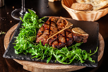 The concept of Italian cuisine. Juicy T-bone steak of beef aging stewed meat, with rucola salad and cherry tomatoes. A glass of red wine and bread basket on the table