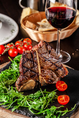 The concept of Italian cuisine. Juicy T-bone steak of beef aging stewed meat, with rucola salad and cherry tomatoes. A glass of red wine and bread basket on the table