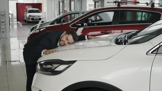 Happy funny man hugging a car that he just bought, in a car dealership / auto shop