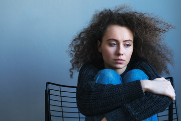 Dark hair teenager with depression sitting alone on the armchair in gray room. Sadness, nostlagic,...