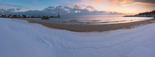 Panorama of winter seascape. The sea coast with snow and cubic stone breakwater with small lighthouse on the edge. Beautiful evening cloudy sky. Long exposure.