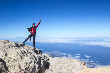 Happy European photographer with camera and tripod standing on mountain top with hand up