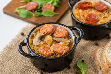 Slovak Christmas national cabbage soup in two small black pots with sausage on the tablecloth background.