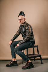 Funny portrait of young stylish man in fashionable clothes sitting on wooden chair with black pot on head in studio on brown background. Handsome smiling boy in glasses, leather jacket, brogue shoes.