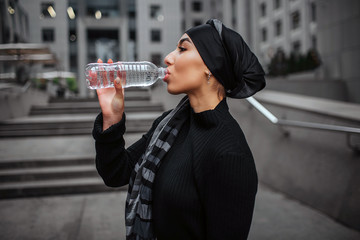 Side shot of young arabian woman drinking water from plastic bottle. She wears black dress and hiijab.