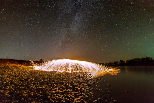 Light painting art concept. Spinning steel wool in abstract circle, firework showers of bright yellow glowing sparkles in fountain form on river bank on blue night starry sky copy space background.