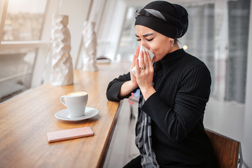 Sick young arabian woman sit inside in cafe. She sneezes in white napkin. Woman suffer. There are cup of coffee and phone at table.