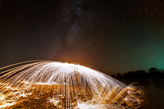 Light painting art concept. Spinning steel wool in abstract circle, firework showers of bright yellow glowing sparkles in fountain form on river bank on blue night starry sky copy space background.