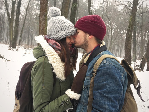 Happy hipster couple in stylish outfits embracing and kissing in snowy winter park. Cute family taking selfie. Atmospheric cozy winter moments. Moody image