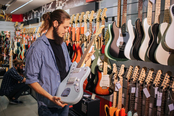 Young bearded musician hold light-blue electric guitar in hands. He looks at it. - obrazy, fototapety, plakaty