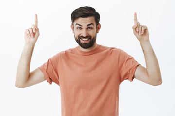 Fototapeta premium Pleased friendly-looking handsome husband in 30s with beard raising eyebrows as pointing up and smiling delighted, suggesting cool copy space, offering great offer over gray background