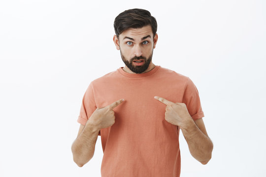 Pissed and questioned adult handsome man being insulted pointing at himself looking confused from under forehead looking at camera bothered and shocked from received offence over gray wall