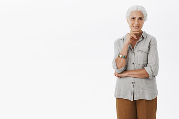 Portrait of charming beautiful old woman with grey combed hair holding hand on chin and gazing interested at camera hearing curious and intriguing promotion deciding how invest money