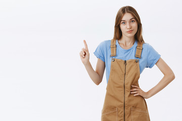 Nah I forbid. Bossy confident and displeased cute female employee in brown overalls and blue t-shirt holding hand on waist shaking index finger in stop or prohibition gesture over grey wall