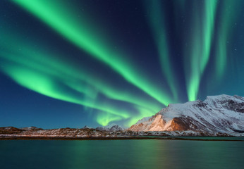 Aurora borealis above the snow covered mountain in Lofoten islands, Norway. Northern lights in winter. Night landscape with polar lights, snowy rocks, reflection in the sea. Starry sky with aurora - obrazy, fototapety, plakaty