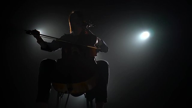 Silhouette of a musician with a cello in a dark studio with lanterns. Black smoke background