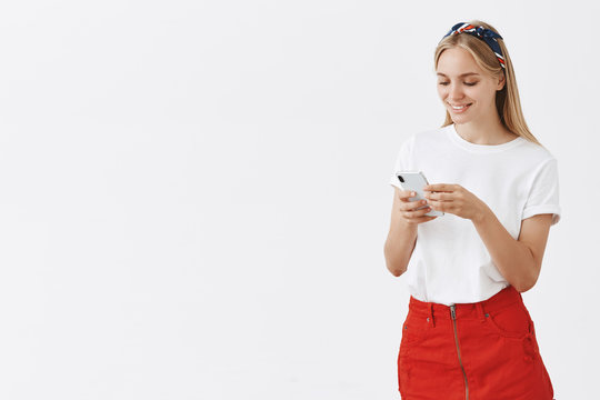 Studio shot of creative and stylish attractive woman with blond hair in headband and red skirt standing half-turned looking at smartphone screen while picking emoji, sending messages
