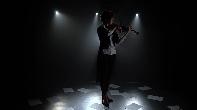 Dark studio girl playing the violin on the floor sheets of notes. Silhouette. Black smoke background