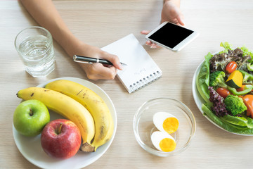 Dieting and calories control for wellness. Woman using smartphone calculate calories of food in breakfast during dieting for lose weight program and take notes. - obrazy, fototapety, plakaty