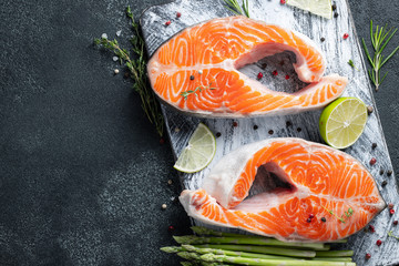 Two raw fresh salmon or trout steaks on ice, rich in omega-3 oil, with lime, thyme and young asparagus on a dark background. Healthy and dietary food. Top view with copy space. Flat lay