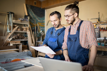 Waist up portrait of two modern carpenters looking at plans while making furniture in joinery...