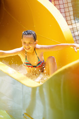 Young pretty girl in a striped colorful swimsuit laughs, rides on a yellow water slide in aqua park.