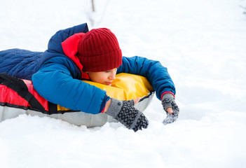 Fototapeta na wymiar cute young boy in hat red scarf and blue jacket lays on tube on snow, has fun, smiles. Teenager rises his hands up in winter park. Active lifestyle, winter activity, outdoor winter games, snowballs