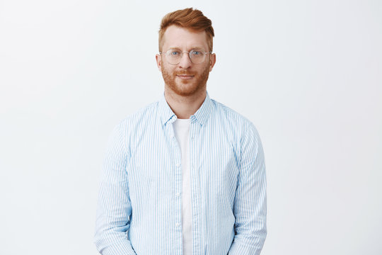 Good-looking creative and smart redhead male entrepreneur hiring new employees, standing in casual shirt and glasses over gray wall, smiling friendly, gazing at camera with thoughtful, determined look