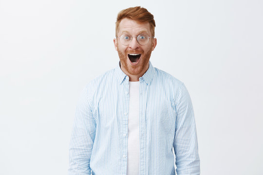 Friends bought me new car, wow. Amazed and thrilled joyful redhead man with beard in glasses and shirt, dropping jaw from amazement and happiness, smiling broadly and gazing at camera fascinated
