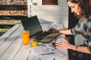 Young woman sitting on outdoor terrace and using laptop and mobile phone. Working mother has freelance job from home. She looking at smartphone screen. Mobile technologies in modern life. Home office.
