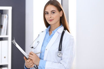 Doctor woman filling up medical form while standing near window in hospital office. Happy physician at work. Medicine and health care concept