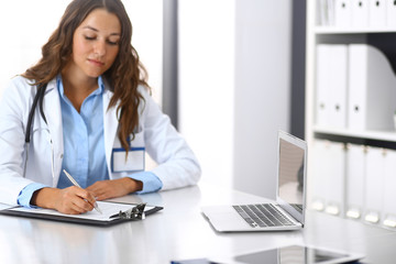 Unknown doctor woman filling up medical form while sitting at the desk in hospital office. Physician at work. Medicine and health care concept