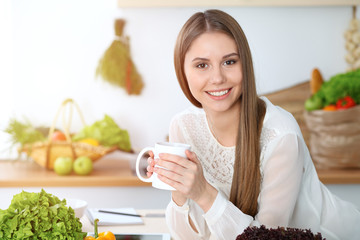 Young happy woman holding white cup and looking at the camera while sitting at wooden table in the kitchen among green vegetables. Good morning, lifestyle or cooking concept