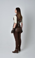 full length portrait of brunette  girl wearing brown leather steampunk outfit. standing pose with back to the camera, on grey studio background.