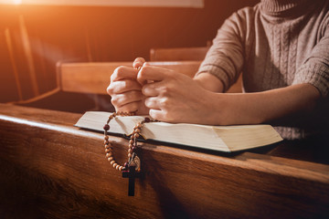 Christian woman praying in church. Hands crossed and Holy Bible on wooden desk.
