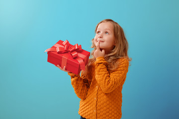 Portrait of a little girl in a warm mustard sweater on a blue isolated background. The child looks dreamily up and holds a red box with a gift.The concept of celebration and receiving a gift