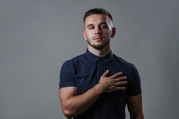 Man swear holding hand to heart. Handsome young man in shirt holding hand on chest on grey