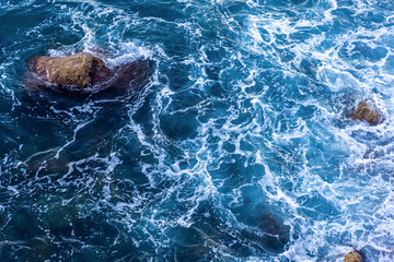 Moving wave and blue water of mediterranean sea touching rock beach making some small white bubble on surface.