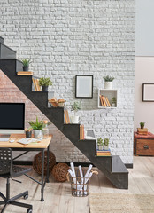 Black stairs in the room, brick wall background, frame book vase of mini plants. Wooden desk and desktop under the stairs.