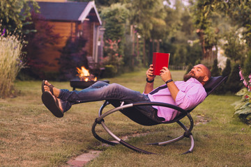 Bearded man reading book on a rocking chair (rocker) in the garden
