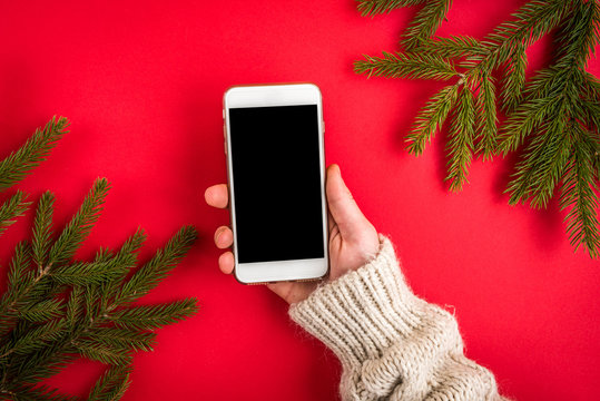 Hand holding mobile phone on red background with Christmas fir branches.