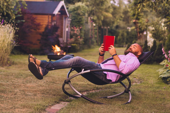 Bearded Man Reading Book On A Rocking Chair (rocker) In The Garden