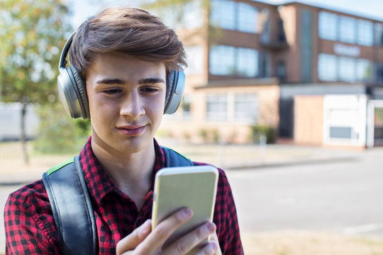 Male Teenage Student Outside College Building Streaming Music From Mobile Phone To Wireless Headphones