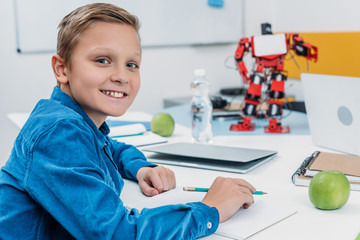 schoolboy sitting at desk with robot model, looking at camera and writing in notebook during STEM lesson