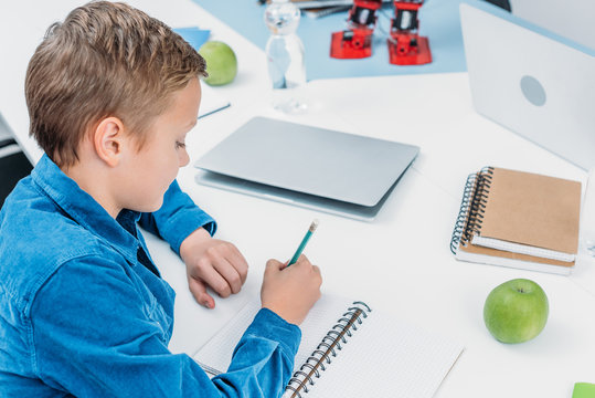 schoolboy sitting at desk and writing on notebook in stem class