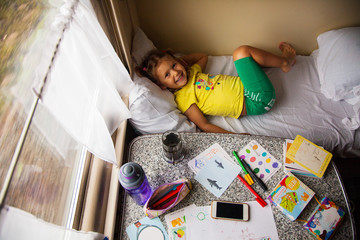 Little girl travelling by train, laying on the fisrt shelf and looking up, top view on the table with travel needs