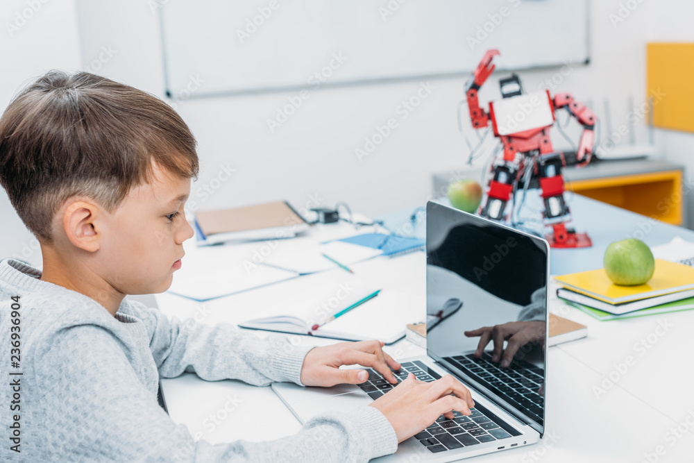 Wall mural concentrated schoolboy typing on laptop keyboard and programming mechanical robot in stem class