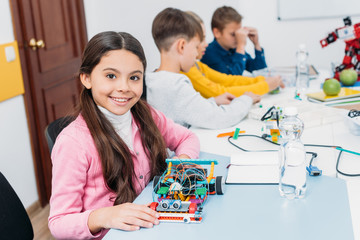 adorable schoolgirl holding robot model, looking at camera while classmates having STEM lesson