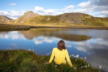 Fototapeta na wymiar A young woman is sitting on the shore of a mountain lake. Clouds of mountains, and blue sky reflected in water. Summer view of travel. Iceland Europe